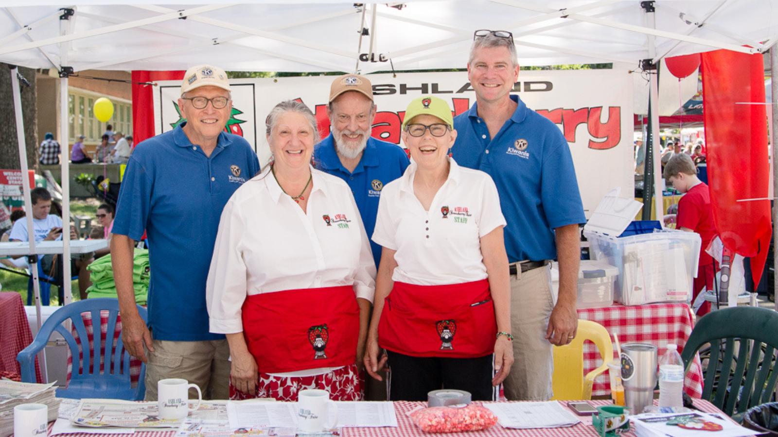 Five volunteers pose under a white canopy at a community fair, wearing branded blue shirts or white shirts with red aprons and badges, smiling for the camera.