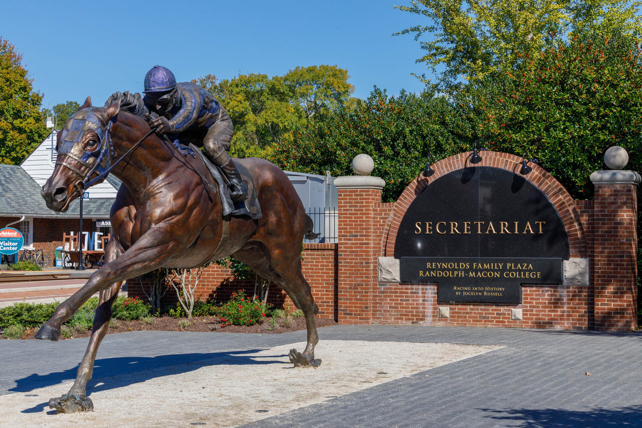 Bronze statue of a jockey on a galloping horse in front of a brick entrance with a sign reading SECRETARIAT and Reynolds Family Plaza, Randolph-Macon College site.