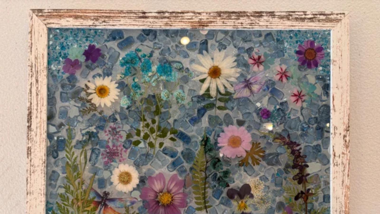 Framed mosaic artwork of colorful wildflowers and daisies on a blue pebble background.