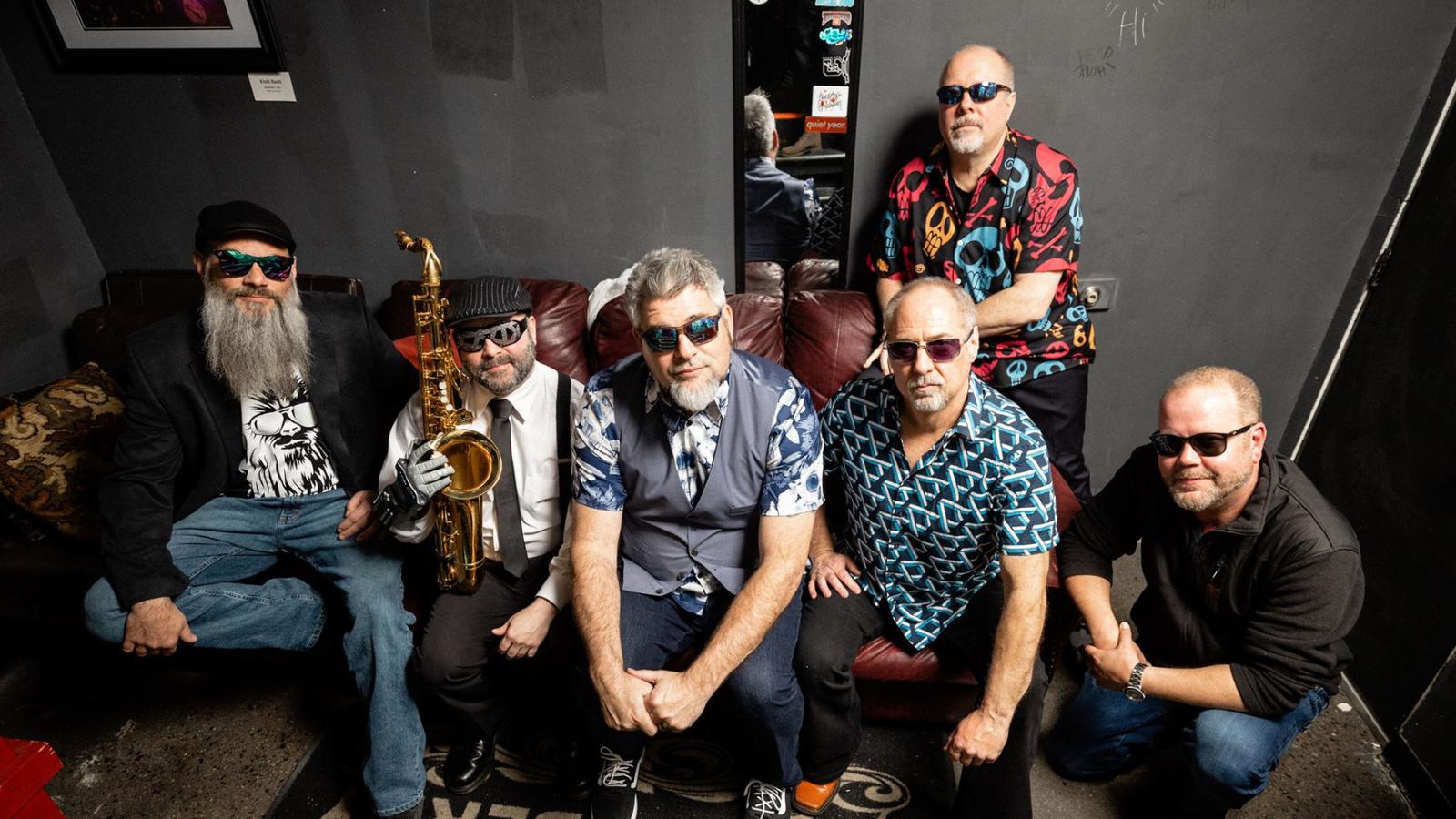 Six men in sunglasses sit and crouch on a leather couch backstage, one with a saxophone, posing for a group photo as a band.