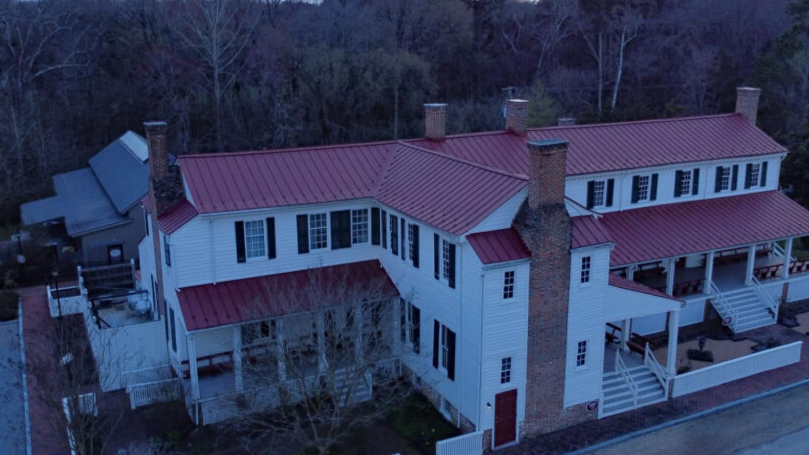 A white two-story house with a red metal roof and brick chimney, set among leafless trees.