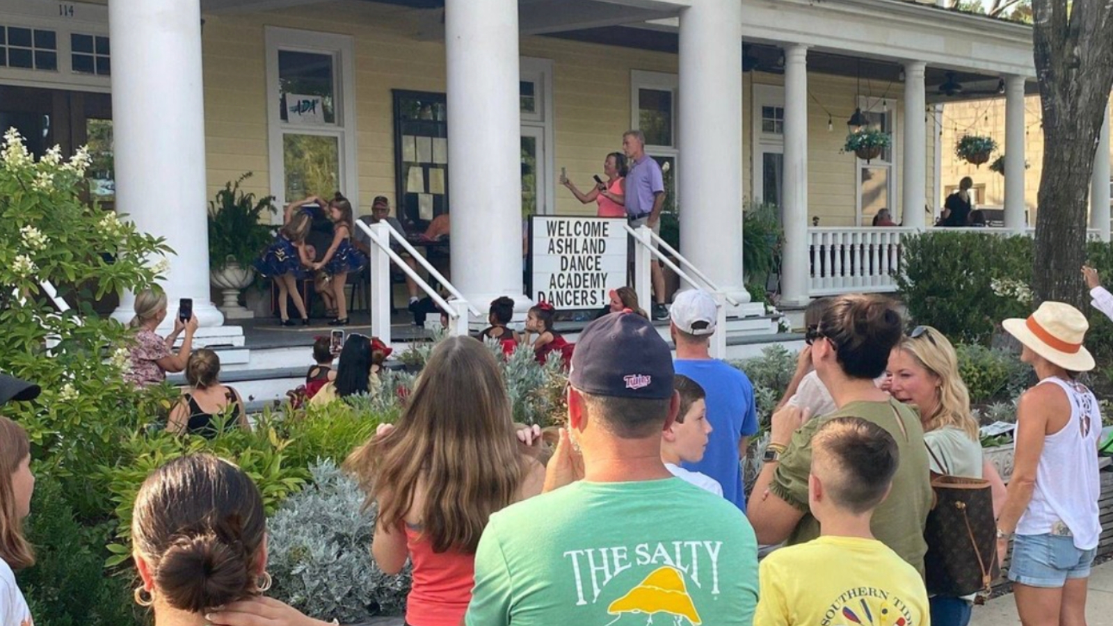 Crowd gathers on a sidewalk in front of a porch as dancers perform on the steps under a sign welcoming Ashland Dance Academy dancers.