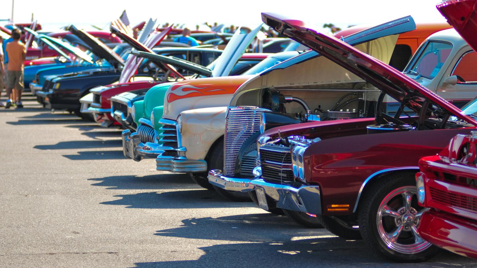 Row of classic cars with open hoods at a car show, showcasing chrome grills and colorful paintwork in the foreground.