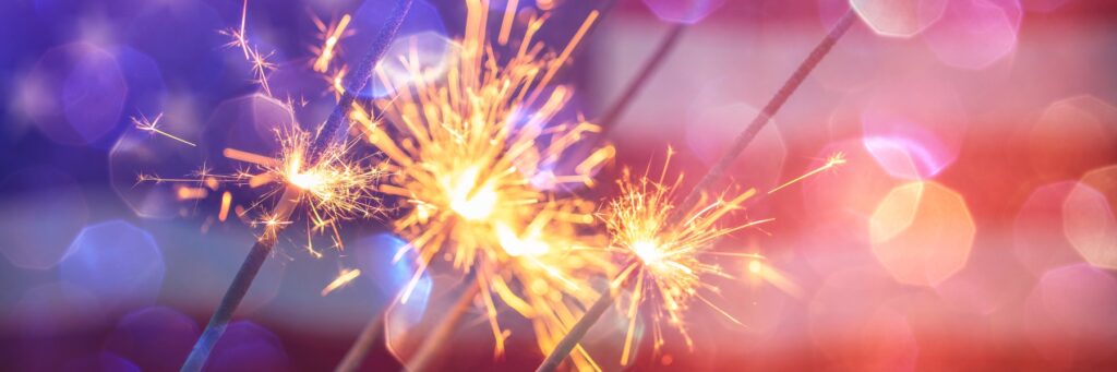 Close-up of sparklers burning, with bright orange sparks against a purple-pink bokeh background, conveying celebration.