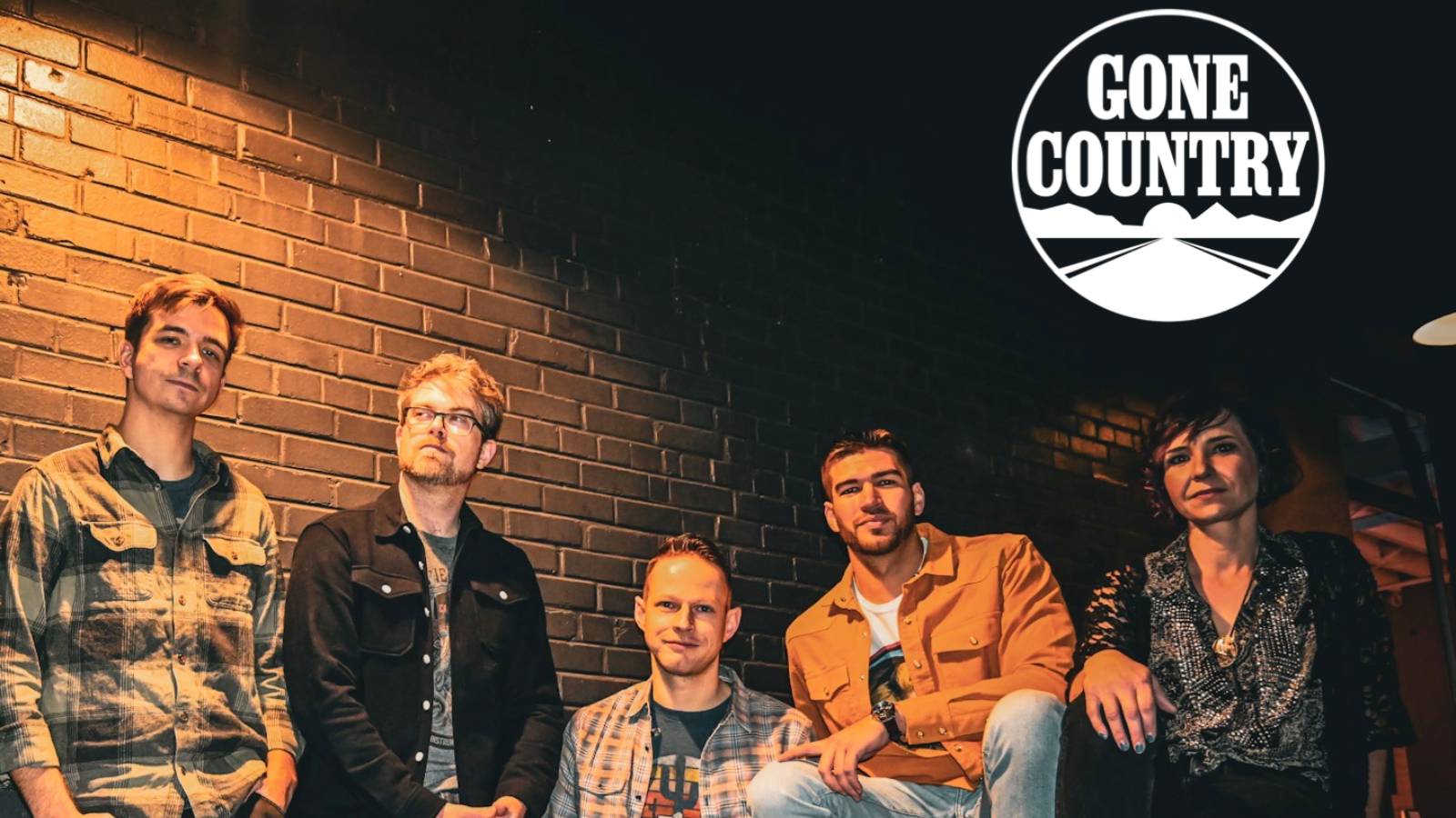 Five people posing against a brick wall at night, with a circular 'Gone Country' logo in the top-right corner.