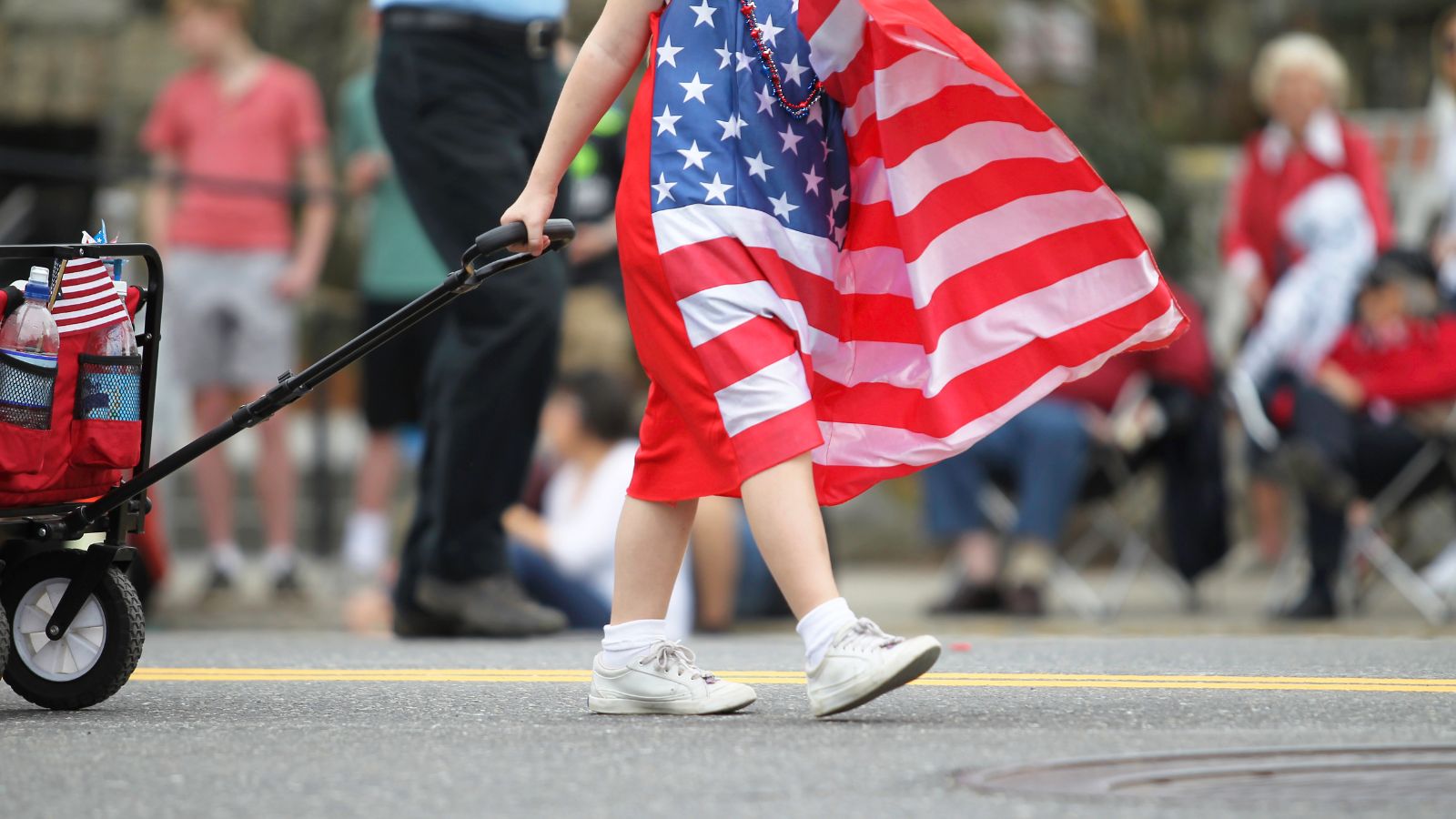 Person wearing a red, white, and blue American flag cape walks down a street at a parade, with a stroller visible on the left and spectators in the background.