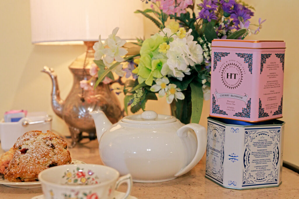 White teapot on a marble counter with a pastry and floral bouquet in the background; pink decorative tea tins to the right.