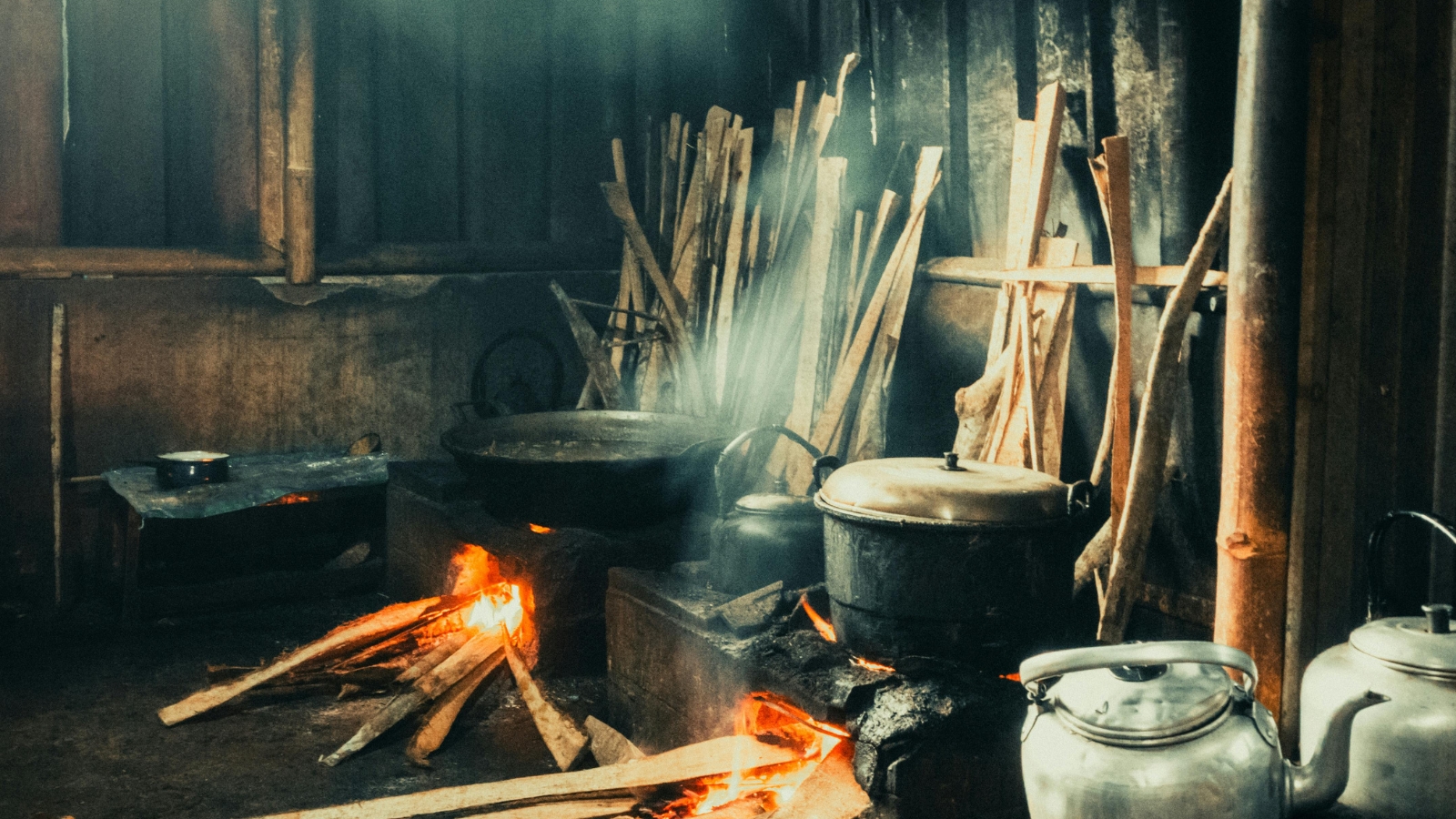 Rustic wood-fired kitchen with a large pot over an open flame and stacked firewood nearby.