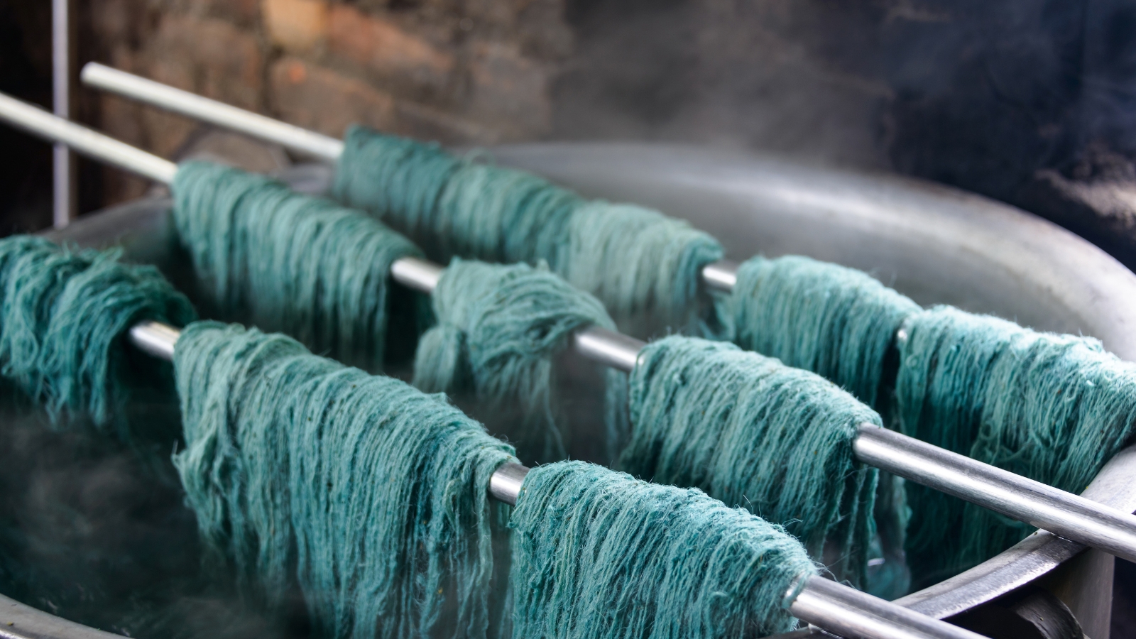 Teal yarn skeins draped over metal bars inside a steaming dye vat in a textile mill