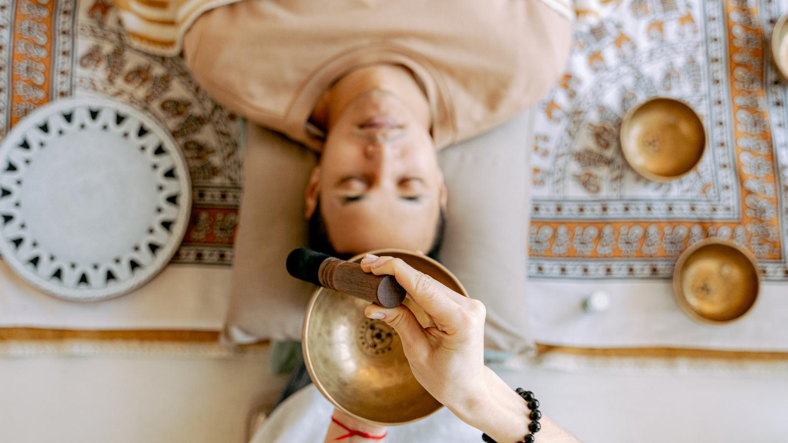 Person lying on a cushion while a practitioner taps a brass singing bowl during a sound healing session on a patterned rug.