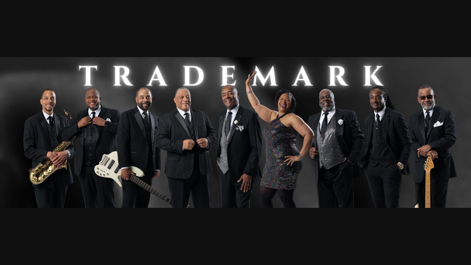 Nine musicians in black suits stand together against a dark backdrop with the word TRADEMARK above them, holding saxophone, bass guitar, and electric guitar.