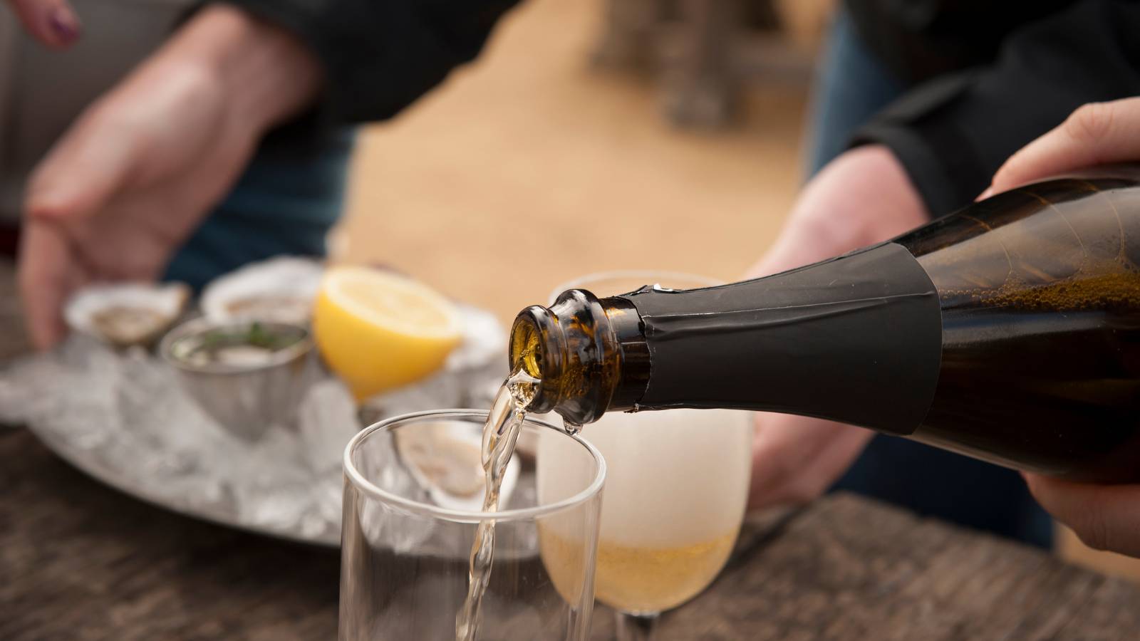 Person pours sparkling wine from a dark bottle into a glass as ice and a lemon half sit on a tray in the background, suggesting a festive outdoor setting.